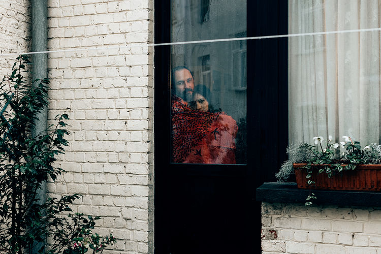 Couple wrapped in a red blanket looking outside from a backyard door window on a grey rainy day