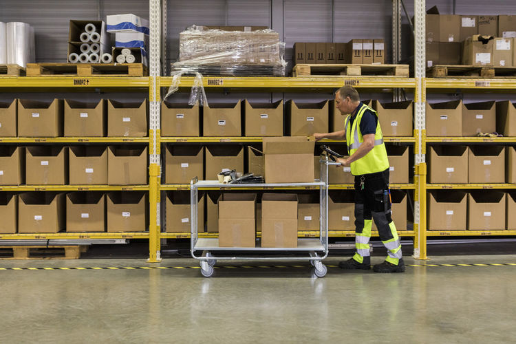 Full length side view of senior male worker pushing cart with cardboard boxes by rack at distribution warehouse