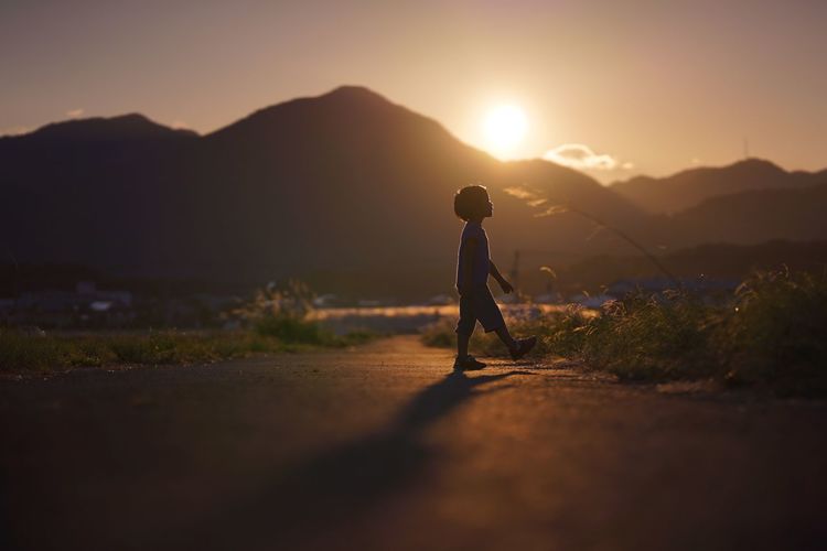 Silhouette boy walking on road towards mountain against sky during sunset