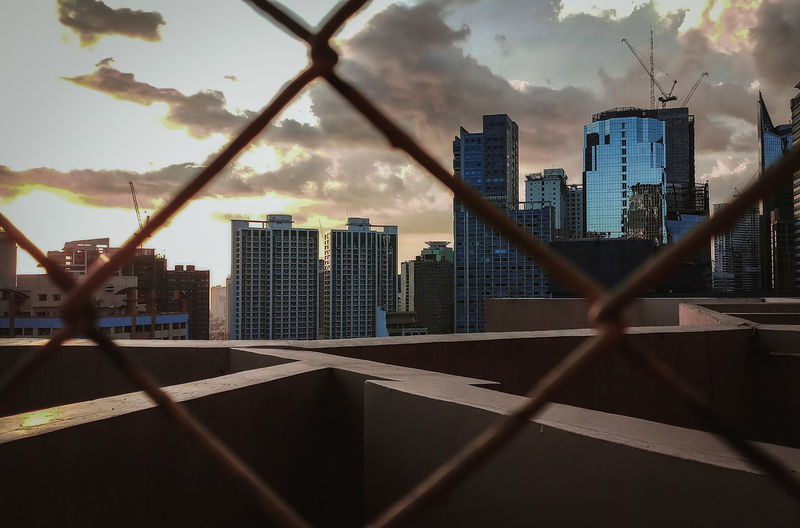Modern buildings against sky seen through chainlink fence at sunset