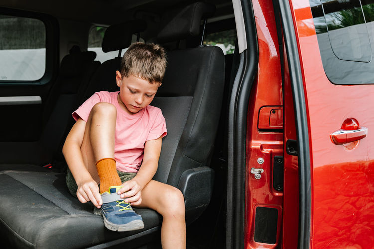 Boy in casual clothes fastening shoe while sitting on back seat of red vehicle during road trip
