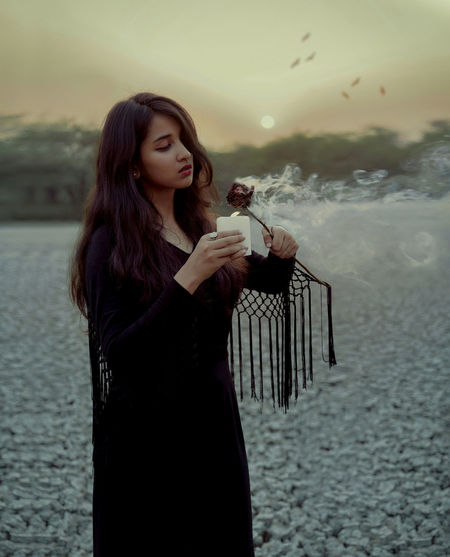 Young woman burning rose with candle while standing outdoors