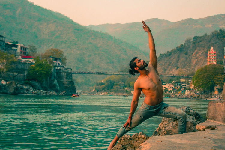 Shirtless man practicing yoga by river against mountains