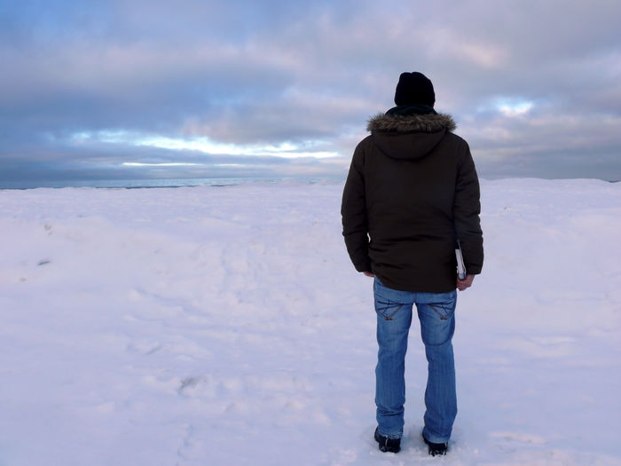 Full length rear view of man standing on snow covered field against cloudy sky