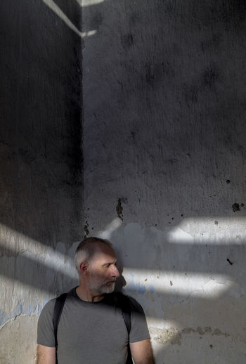 Portrait of adult man looking away while standing on old wall with sunlight and shadow