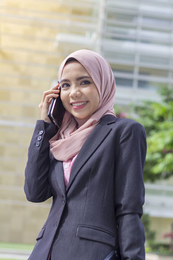 Low angle portrait of happy businesswoman using smart phone while standing against office building
