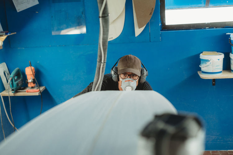 Craftsman in protective mask and headphones making surf board in small workshop with blue walls