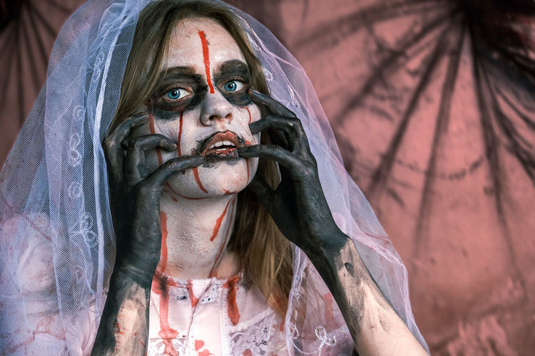 Close-up portrait of zombie woman in wedding dress with veil and stage makeup looking into camera. 
