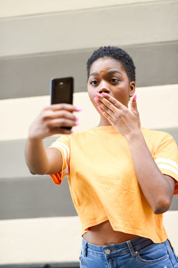 Shocked young woman taking selfie with mobile phone against wall