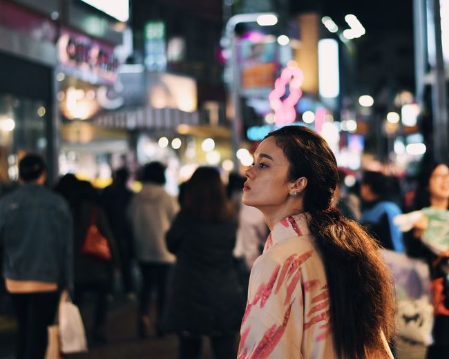 Woman looking away while standing in illuminated city at night