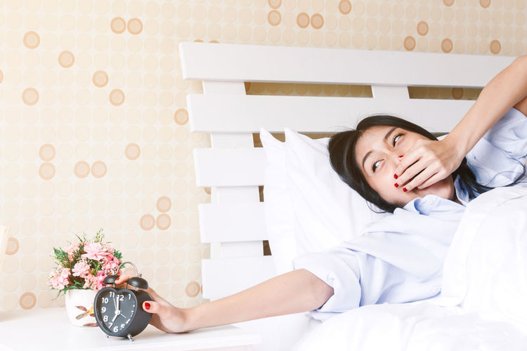 Yawning woman holding alarm clock while lying down on bed