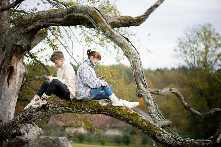 Mother and son teen sit with their backs together on a tree branch and use cell phones and headsets 