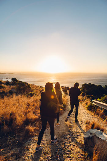 Rear view of friends walking at beach against clear sky during sunset