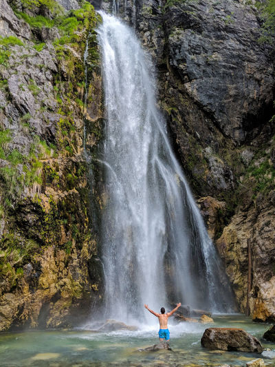 Rear view of shirtless young man with arms raised looking at waterfall