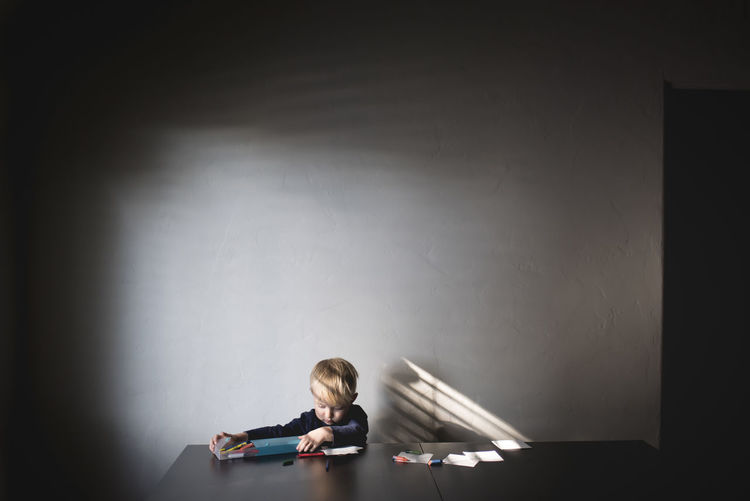 Boy removing crayons from box while sitting at table against wall