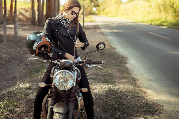 Beautiful woman zipping leather jacket while sitting on motorcycle at roadside