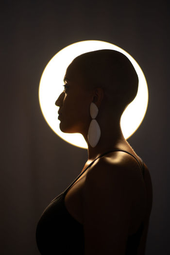 Side view of calm african american female with bald haircut and in trendy earrings standing in dark studio on background of round glowing lamp