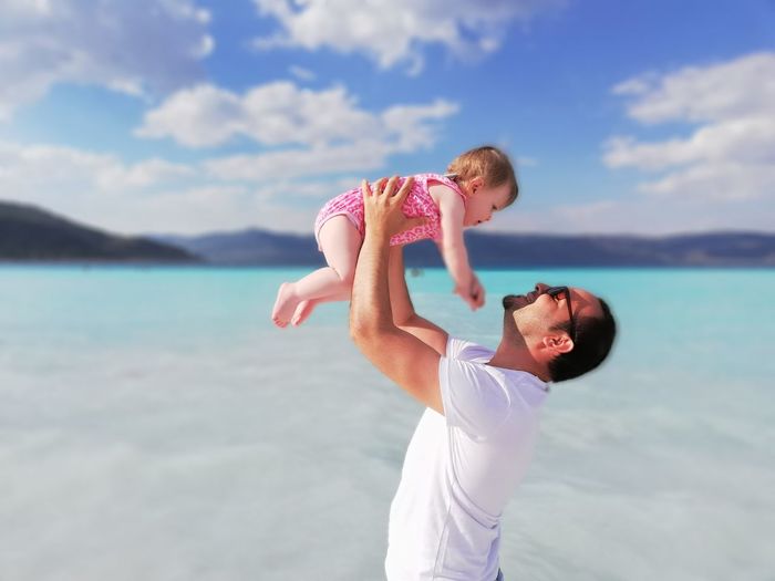 Side view of man picking daughter at beach against cloudy sky