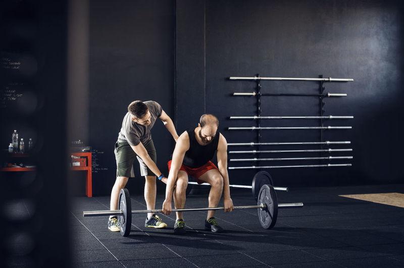 Trainer assisting male athlete in lifting barbell at gym