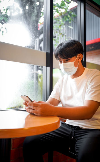 A handsome young teenage boy wear mask playing on smart mobile phone waiting in cafeteria. 