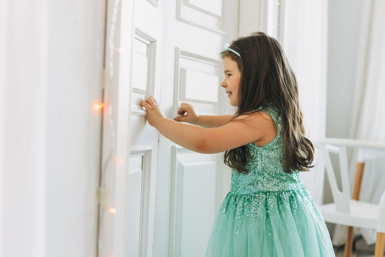Cute happy little girl in mint fancy dress open door at bright room, birthday, christmas time