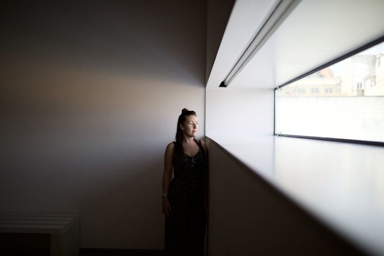 Thoughtful woman looking through window while standing against wall at home