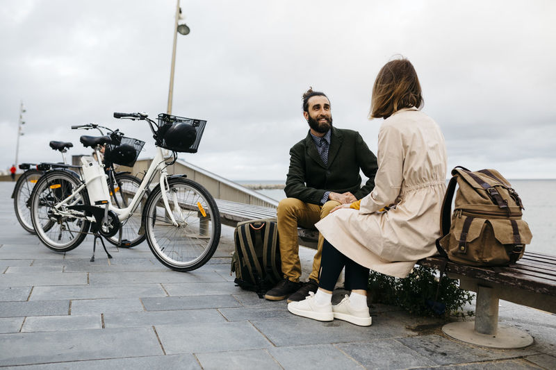 Couple sitting on a bench at beach promenade next to e-bikes talking