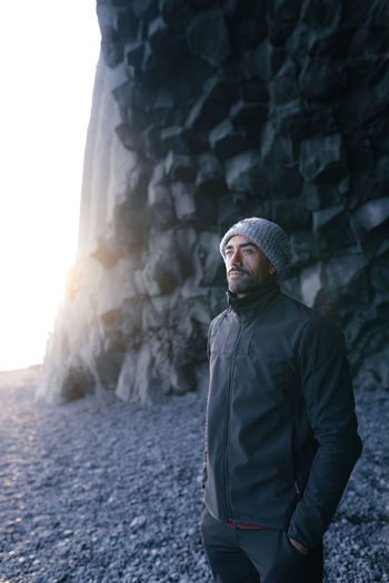 Content young unshaven ethnic male traveler in warm clothes and hat standing near basalt columns on reynisfjara beach with hand in pockets and admiring nature during holiday in iceland