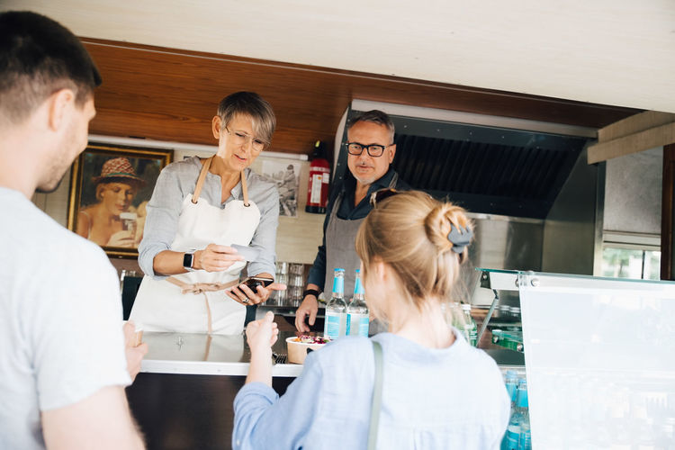 Mature owners serving food to customers while standing with credit card and mobile phone in commercial land vehicle