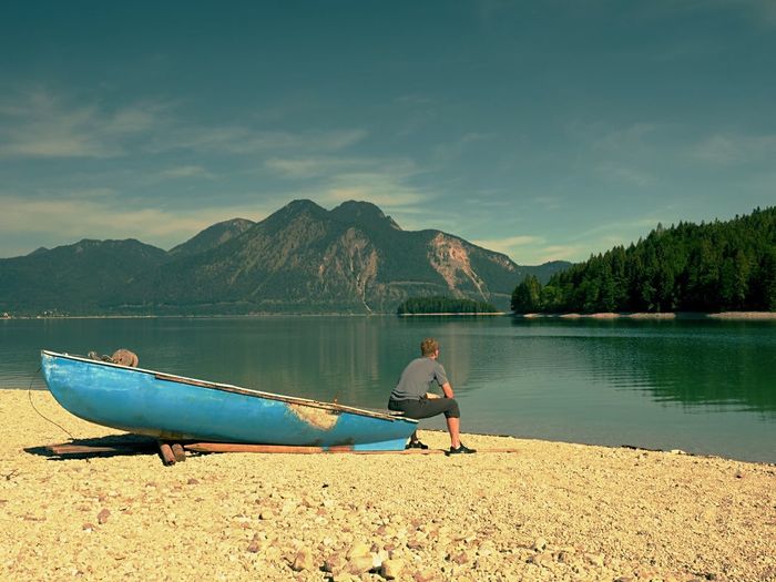 Adult man in blue shirt at old fishing paddle boat at mountains lake coast. sunny spring day.