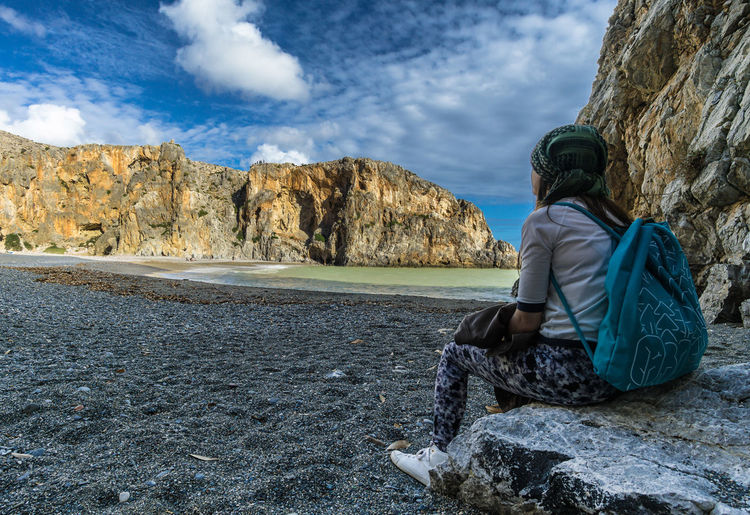 Rear view of woman sitting on rock by sea against sky