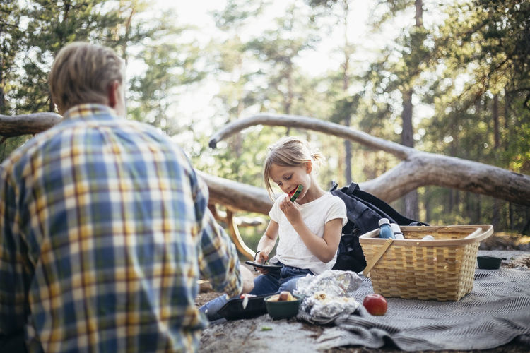 Daughter eating watermelon while sitting with father on picnic blanket in forest