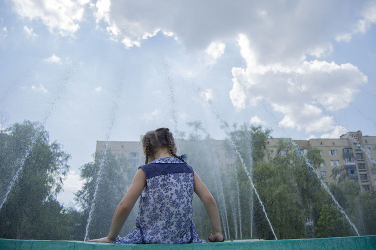 Rear view of girl sitting by fountain against sky