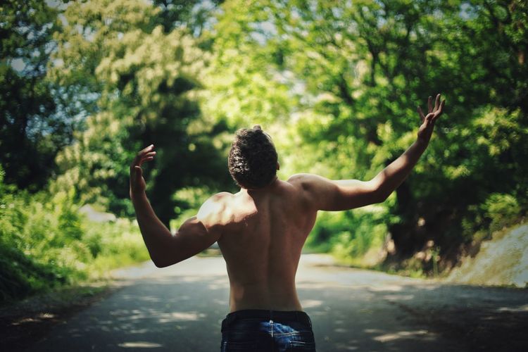 Rear view of shirtless man gesturing while standing on road amidst trees