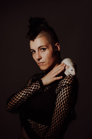 Confident woman with mohawk carrying white rat on shoulder and looking at camera against black background