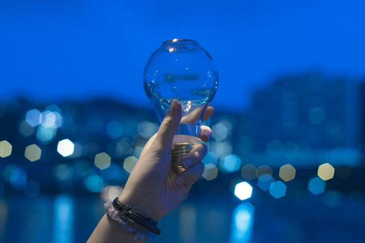 Cropped hand of woman holding light bulb with water against sky