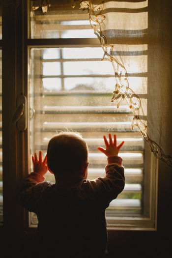 A baby looking through a window covered with lace curtain in sunset
