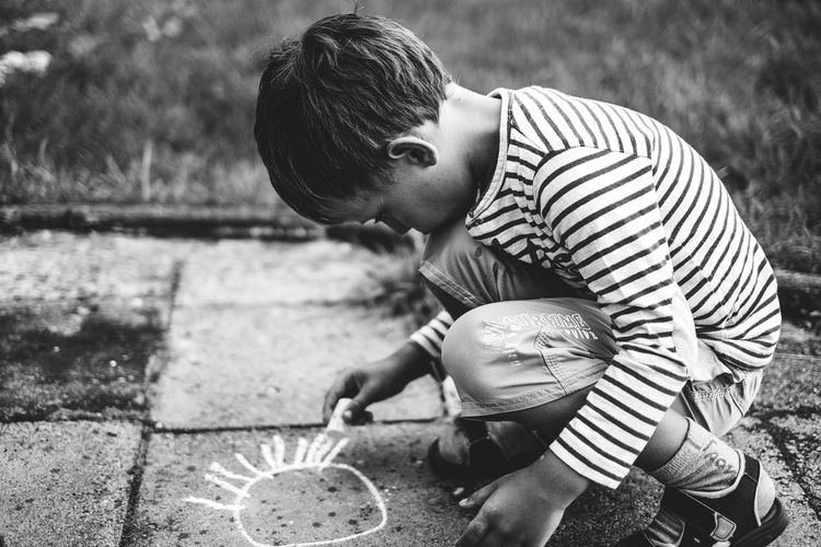 Side view of boy chalk drawing while crouching on street