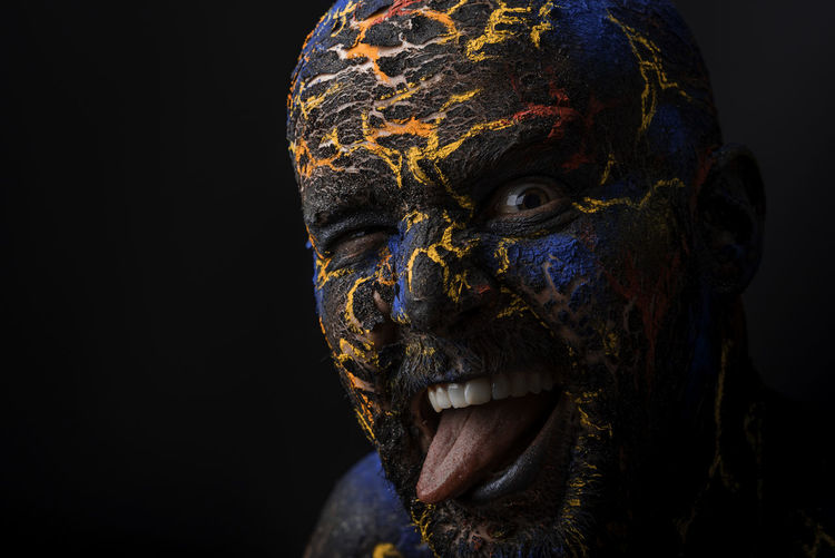 Close-up portrait of mid adult man with body paint sticking out tongue standing against black background