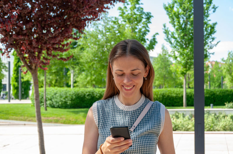 Young woman using phone while standing by trees