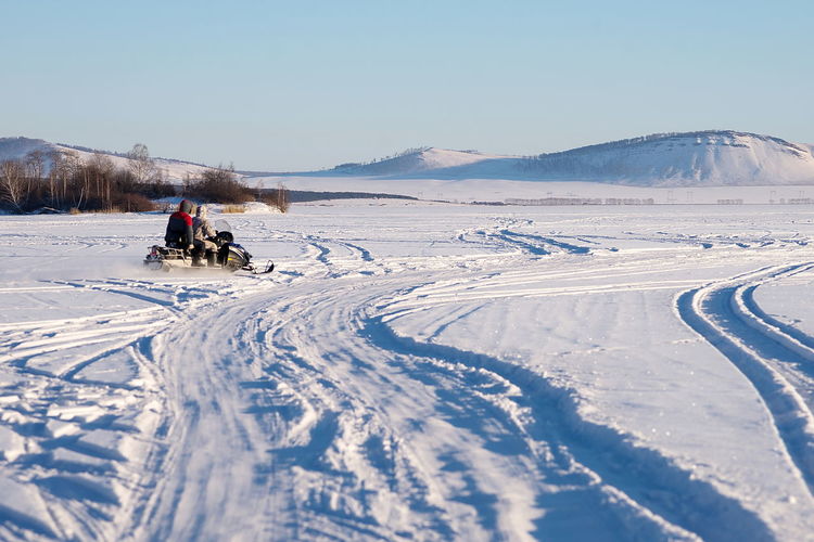 The snowmobile with fishermen is riding along the winter lake against the backdrop of the mountains. 