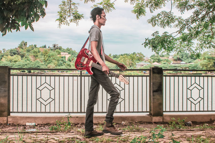 Young man holding guitar standing against trees
