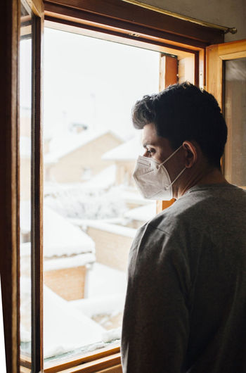 Young man with mask in quarantine looking at the snowfall through the window of his house