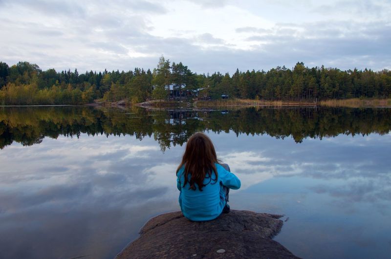 Rear view of girl looking at lake while sitting on rock in forest