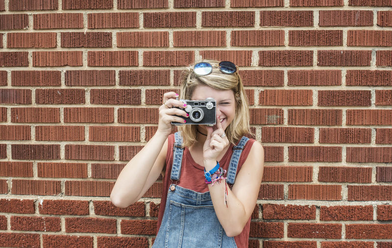 Portrait of young woman photographing against brick wall