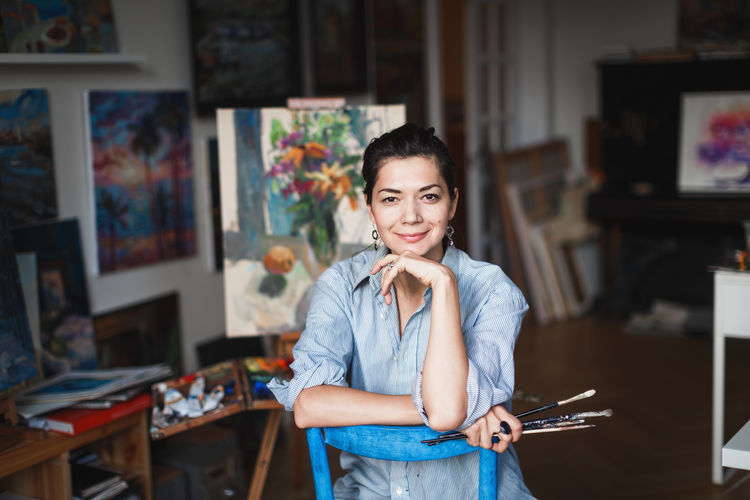A young smiling brunette woman artist in her Studio is holding a brush. Near her easel, paintings and various art equipment. Artist Studio Art And Craft Beautiful Woman Brush Casual Clothing Creativity Emotion Focus On Foreground Front View Happiness Indoors Looking At Camera One Person Painter Portrait Real People Smiling Standing Table Young Adult Young Women International Women's Day 2019