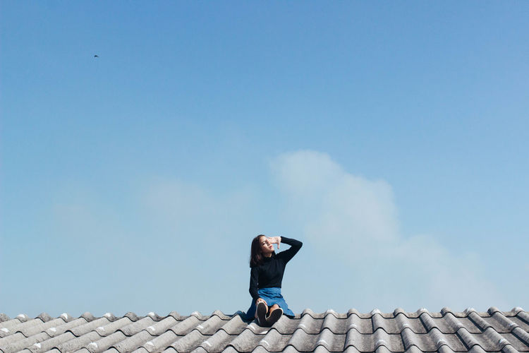 Woman shielding eyes while sitting on rooftop against sky