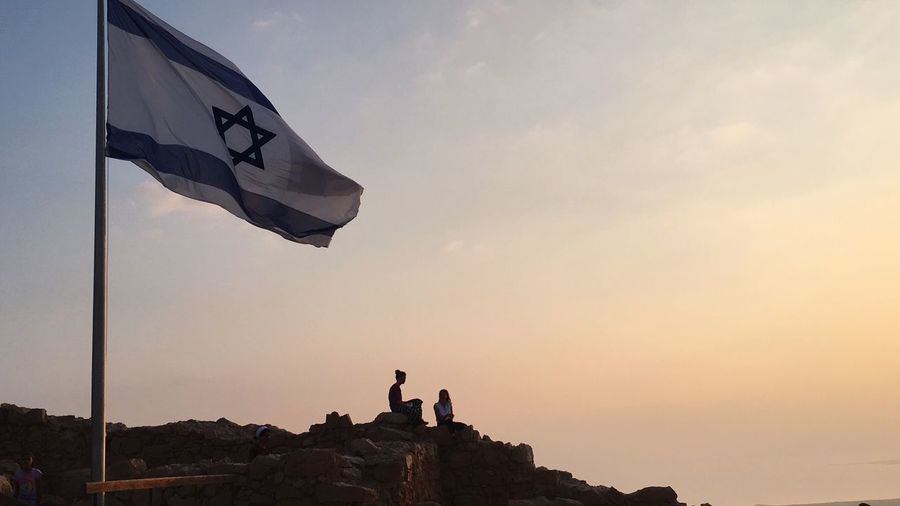 People on rock against sky during sunrise on top of mount masada, next to israeli flag