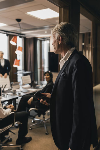 Senior male professional standing on doorway looking at colleagues during meeting board room