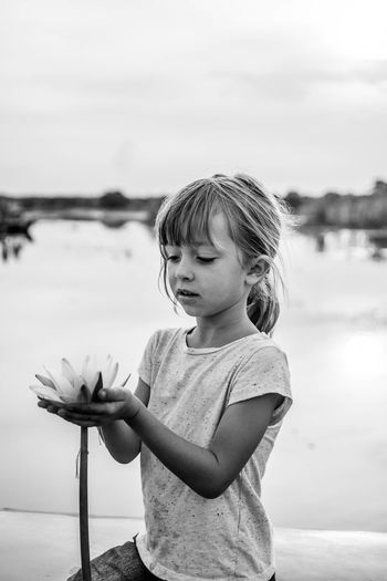 Cute girl holding lotus while sitting by lake against sky